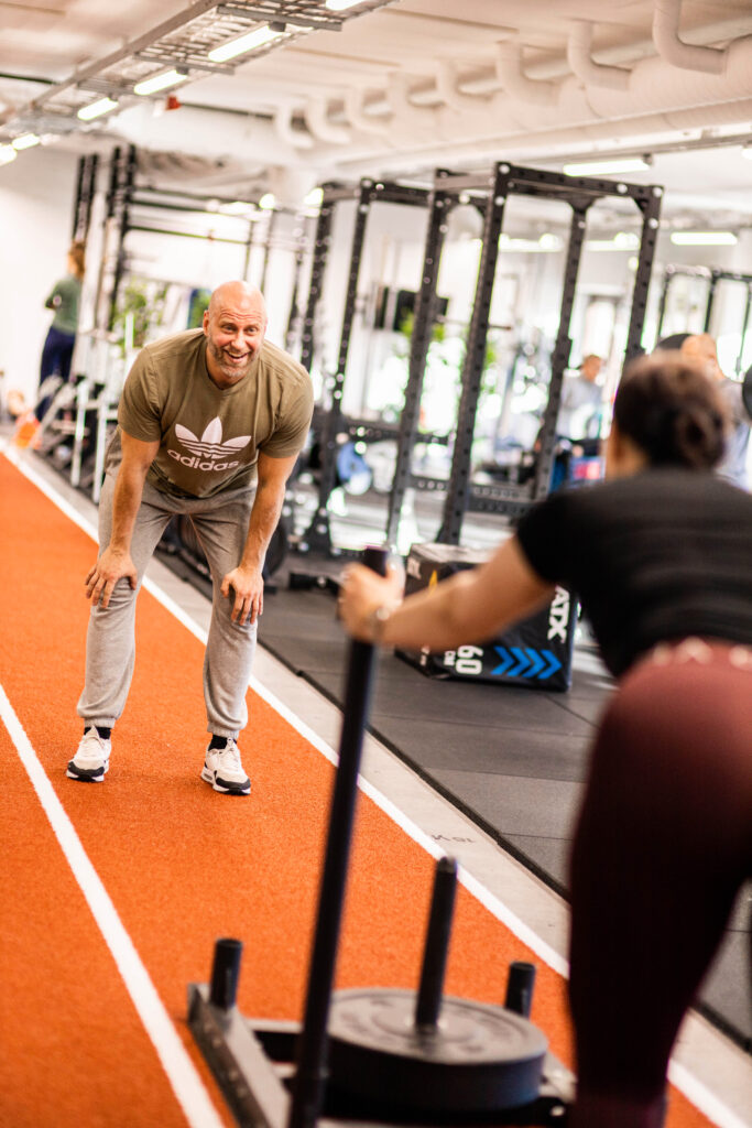 Nainen työntää kelkkaa kuntosalilla Metsälässä. Osteopaatti seisoo hänen edessään, nojaa polviinsa ja katsoo naista hymyillen.